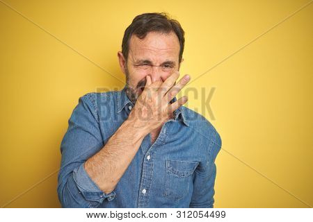 Handsome middle age senior man with grey hair over isolated yellow background smelling something stinky and disgusting, intolerable smell, holding breath with fingers on nose. Bad smells concept.