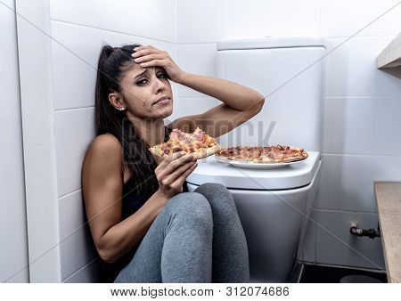 Young Beautiful Bulimic Woman Sitting On The Bathroom Floor Eating Pizza Looking Guilty