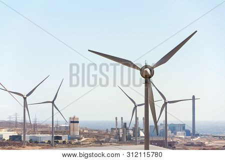 White Turbines At Wind Farm With Power Station And Sea In Background, Arid Landscape On Sunny Summer