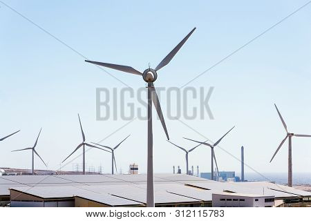 White Turbines At Wind Farm With Power Station And Sea In Background, Arid Landscape On Sunny Summer
