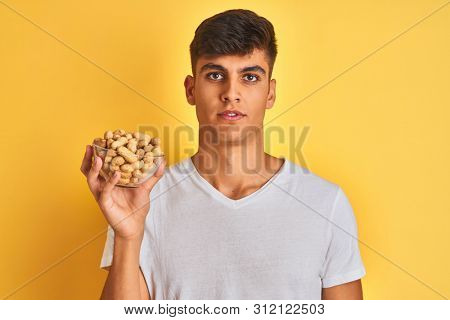 Young indian man holding bowl with peanuts over isolated yellow background with a confident expression on smart face thinking serious