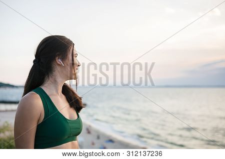 Pretty Brunette Woman Resting After Mornong Jogging At The Sea Shore At Sunrise Listening To The Mus
