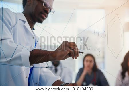 Young Male African Doctor Draws Molecular Formula On Glass Board. A Teacher In A Lab Writing Chemica