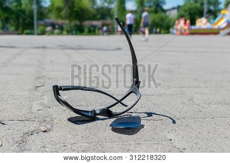 Broken Sun Glasses Lie On A Stone Tiled Walkway In The City Park Against The Background Of Residenti