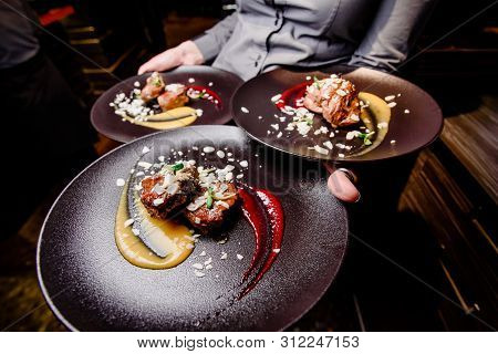 Woman Waiter Carries Three Plates Of Food. Meat Steak Garnished With Two Sauces On A Dark Plate. Clo