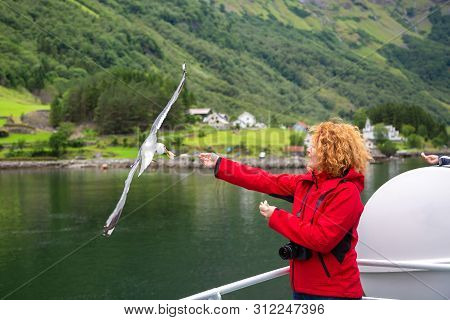 Woman In Red Jacket Traveling On Ferry Boat And Feeding Seagull. Young Woman Feeding Seagulls Flying