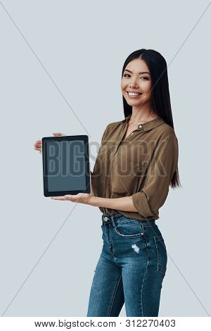 Look Here! Beautiful Young Asian Woman Looking At Camera And Smiling While Standing Against Grey Bac