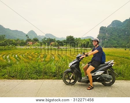 Girl In Helmet On A Scooter Standing On Side Road Looking At Camera With Beautiful Green Rice Fields