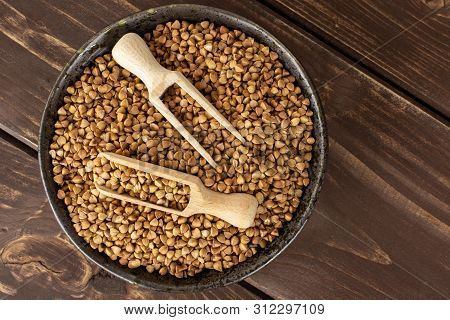 Lot Of Whole Raw Brown Buckwheat Grain In A Grey Ceramic Bowl With Wooden Scoop Flatlay On Brown Woo