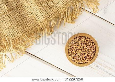 Lot Of Whole Raw Brown Buckwheat Grain In A Wooden Bowl On Jute Cloth Flatlay On White Wood