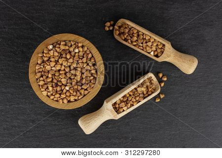 Lot Of Whole Hulled Raw Brown Buckwheat Grain In A Wooden Bowl With Wooden Scoop Flatlay On Grey Sto