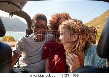 Three happy young adult friends sitting in an open car by the coast, close up, waist up