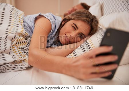 White woman half asleep in bed looking at smartphone, her partner sleeping in background, close up