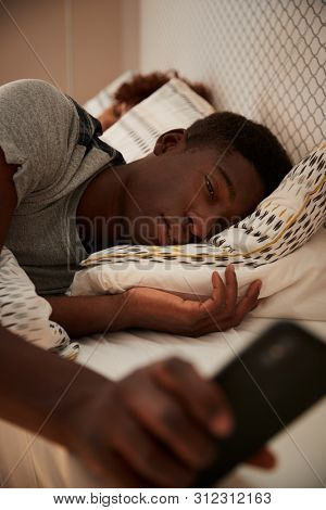 Millennial African American man half asleep in bed holding smartphone, his partner in the background, vertical