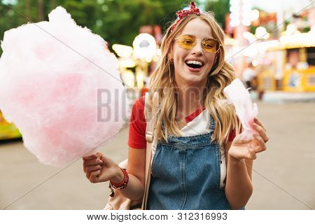 Image of a cheerful laughing young blonde woman in amusement park holding cotton candy candyfloss.