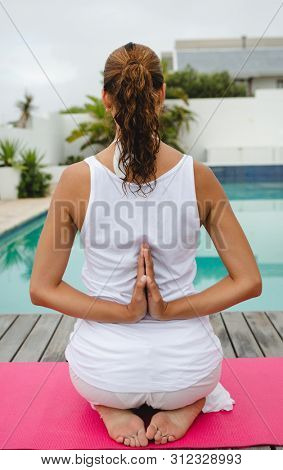Rear view of mixed-race woman performing yoga near swimming pool in the backyard. Summer fun at home by the swimming pool
