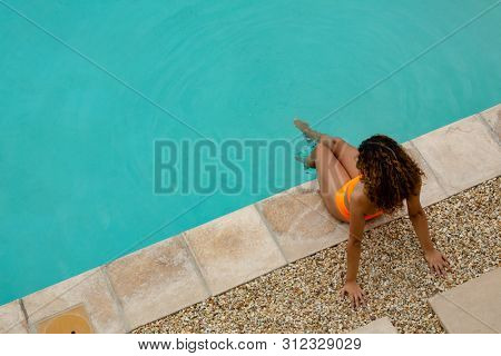 High angle view of mixed-race woman sitting at the edge of swimming pool in the backyard at home. Summer fun at home by the swimming pool