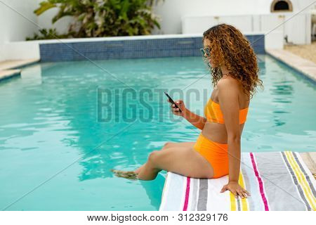 Side view of beautiful mixed-race woman using mobile phone while sitting at the edge of swimming pool. Summer fun at home by the swimming pool