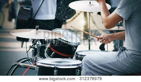 A Guy In Gray Clothes Plays On A Red And Black Drum Set With Wooden Sticks, Speaking At A Presentati