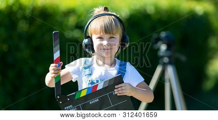 A Little Girl In Headphones With A Tv Numerator In Her Hands Is Standing Next To Camera On A Tripod.