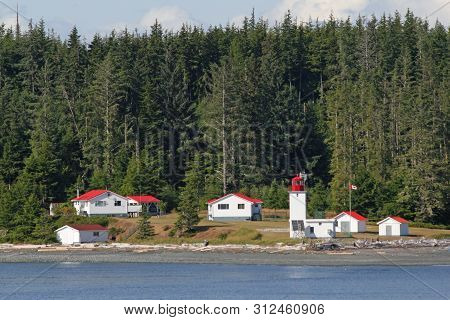 Inside Passage, Canada - August 5, 2006 - Lighthouse And Red-roofed Buildings With Evergreen Forest 