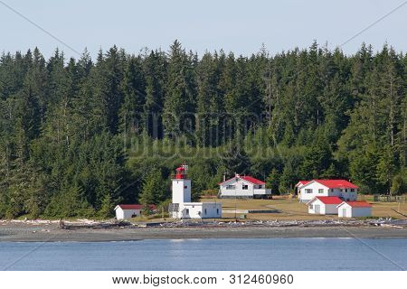 Inside Passage, Canada - August 5, 2006 - Lighthouse And Red-roofed Buildings With Evergreen Forest 