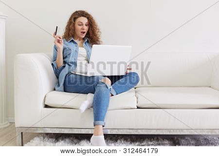 Indoor Shot Of Shocked Emotional Lady With Curly Hair Sitting In Light Living Room, Raising One Hand