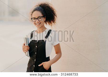 Lovely Curly Woman With Combed Hair, Dressed In White T Shirt, Sarafan, Keeps Hand In Pocket, Holds 
