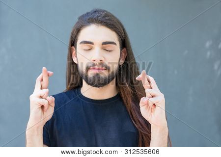 Calm Young Bearded Man With Closed Eyes Holding Cross Fingers. Stylish Brunet With Long Hair And Pie