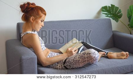 Young Fitness Woman In Sportswear Doing Yoga Stretching Leg While Reading Book On Sofa In Living Roo