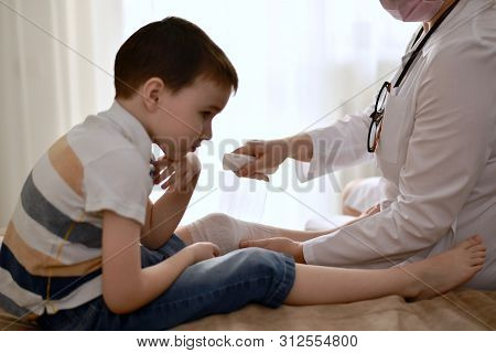 The Hands Of The Doctor Carefully Bandage The Knee Of The Child Who Is Distressed From The Injury.
