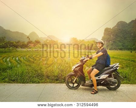 Girl In Helmet On A Scooter Standing On Side Road Looking At Camera With Beautiful Green Rice Fields