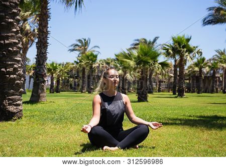 Girl Doing Yoga On The Grass Among Palm Trees In The Fresh Air.