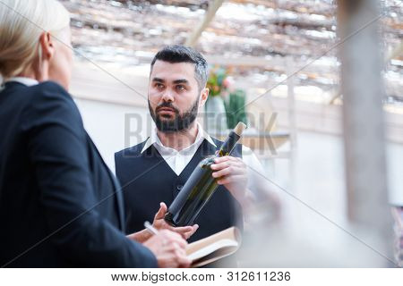 Young confident cavist with bottle of wine talking to his colleague