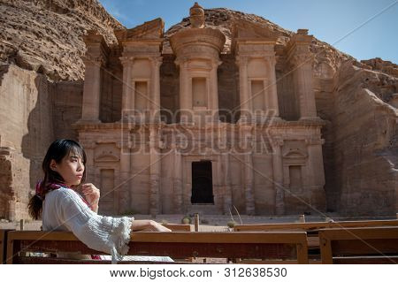Asian Woman Tourist In White Dress Sitting And Looking At Ad Deir Or El Deir, The Monument Carved Ou