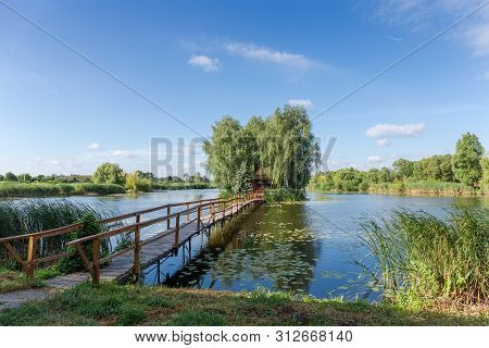 Small Wooden Footbridge To The Tiny Island With Trees And Romantic Fishing Hut On Lake In Summer Mor