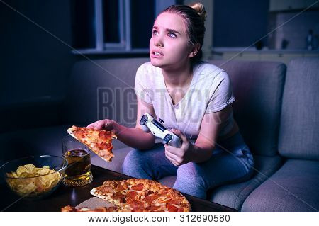 Young Woman Playing Game At Night. Hungry Model Look Up On Screen. Holding Slice Of Pizza And Gamepa