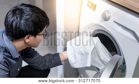 Asian Man Loading White Clothes Into Front Door Washing Machine In Laundry Room. Housework Or Chores