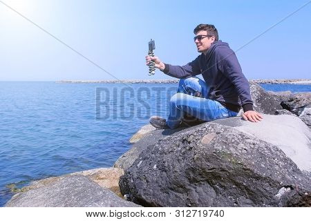 Man Talks Video Chat At Sea On Smartphone With Small Tripod Sits On Stone. Young Guy Traveller Share