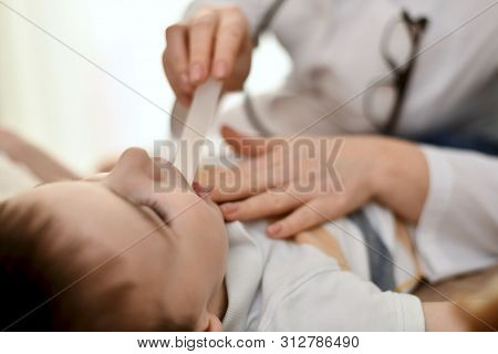 A Doctor With A Spatula Inspects The Child Throat. The Spatula Is In The Patient Mouth.