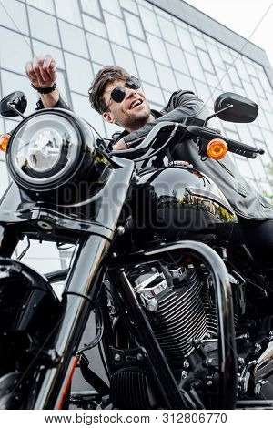 Low Angle View Of Handsome Man In Leather Jacket Sitting On Motorcycle, Amiling And Looking Away