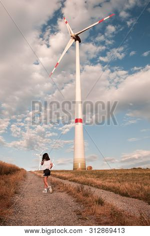 Girl Walking On The Field Of Cereal. Windmill On Background. Innovation And Recouvery Resources Conc
