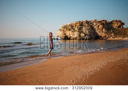 Beautiful Girl In A Dress Walks Along The Sea Sandy Shore At Sunset.