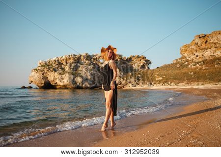 Beautiful Girl In A Black Dress And Hat Walks Along The Sea Sandy Shore At Sunset