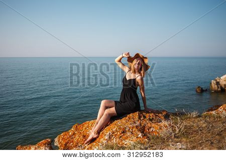 The Girl In A Black Dress And Hat On A Rocky Beach Looks At The Sea