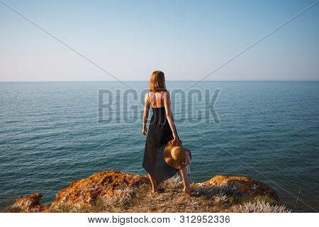 The Girl In A Black Dress And Hat On A Rocky Beach Looks At The Sea