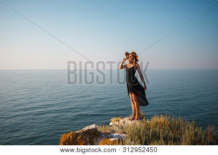 The Girl In A Black Dress And Hat On A Rocky Beach Looks At The Sea