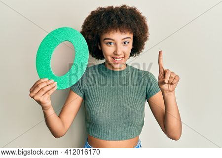 Young hispanic girl holding number zero smiling with an idea or question pointing finger with happy face, number one 