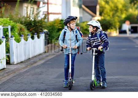Two School Kid Boys In Safety Helmet Riding With Scooter In The City With Backpack On Sunny Day. Hap