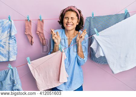Young brunette woman doing laundry around string hangs gesturing finger crossed smiling with hope and eyes closed. luck and superstitious concept. 